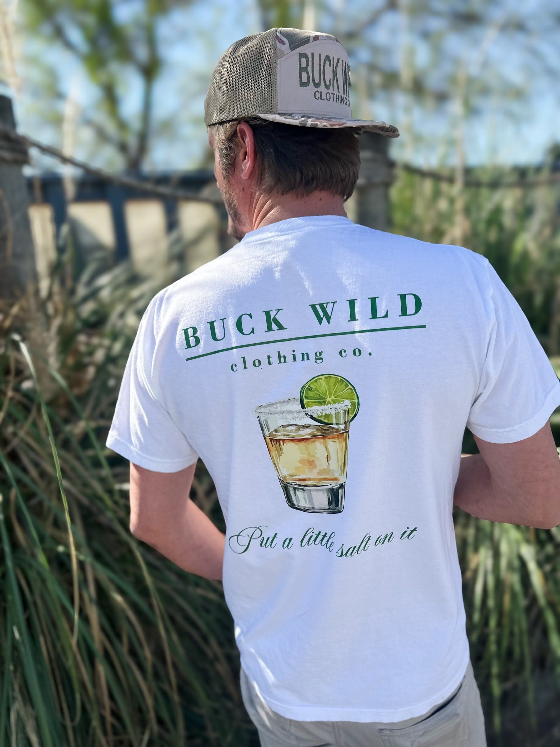 A man in a white Buck Wild Clothing Co. tee and camo hat standing in the marsh grass on a sunny afternoon